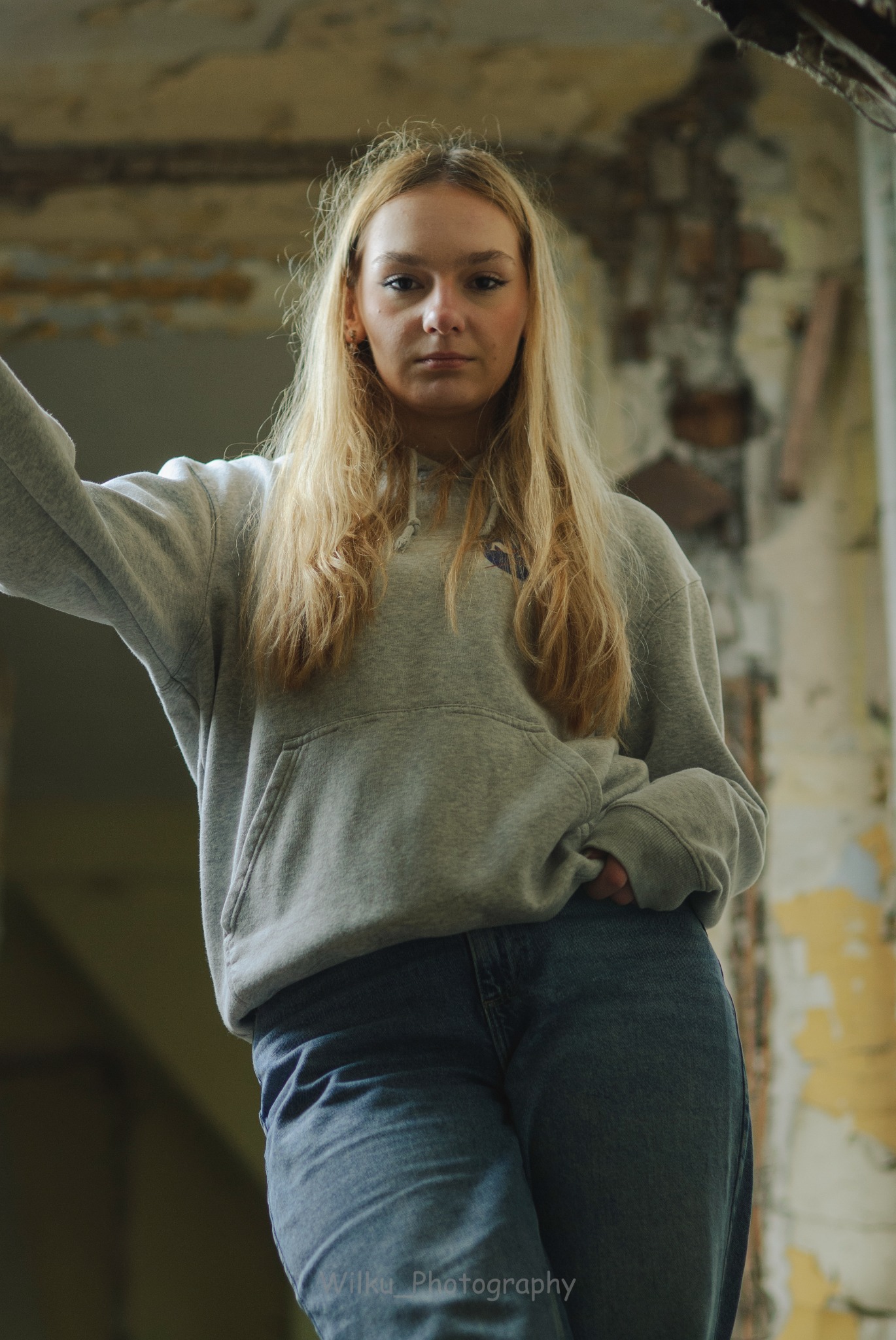 Portret kobiety w bluzie z kapturem i jeansach, stojącej w opuszczonym budynku z odpadającą farbą ze ścian.