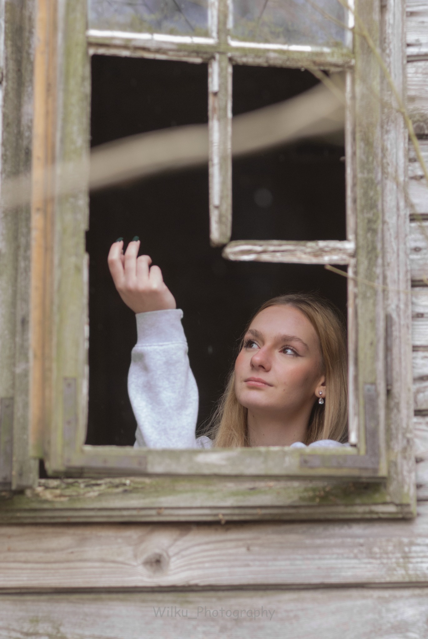 Portret młodej kobiety z blond włosami, spoglądającej w górę przez zniszczone, drewniane okno.
