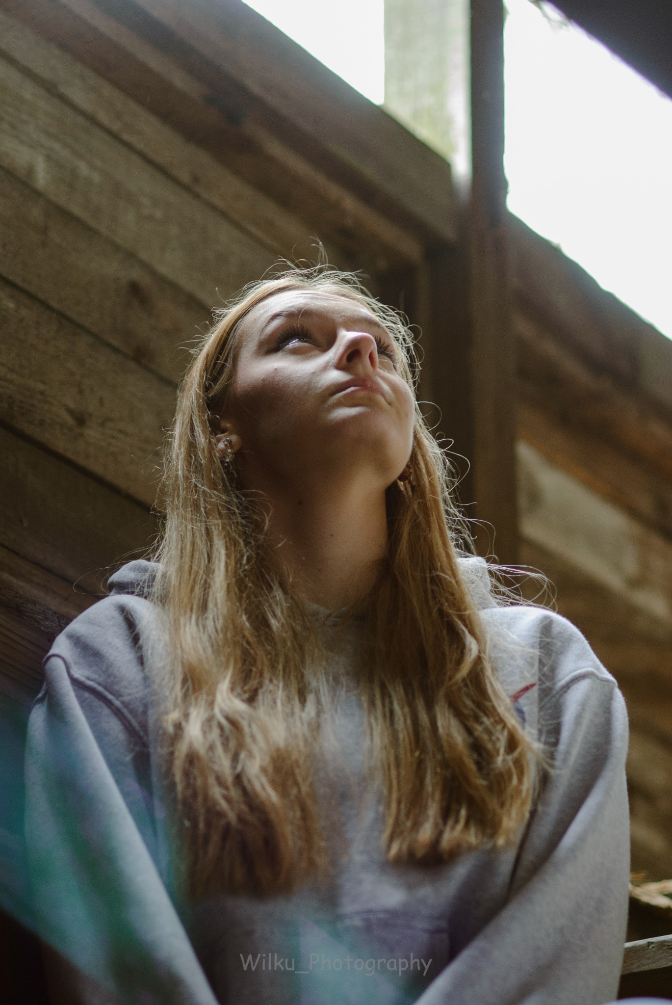 Portret młodej kobiety spoglądającej w górę, z drewnianym tłem i naturalnym oświetleniem z góry.