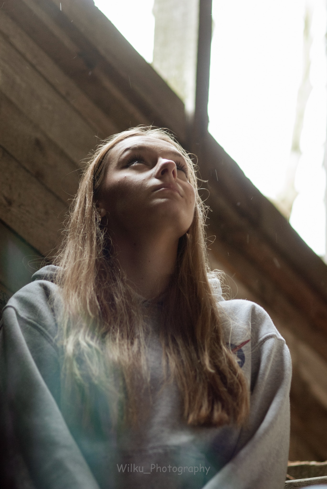 Portret młodej kobiety z długimi blond włosami, ubrana w szarą bluzę z logo NASA, spogląda w górę w kierunku jasnego okna, z drewnianymi elementami w tle i delikatnymi kroplami deszczu w powietrzu.