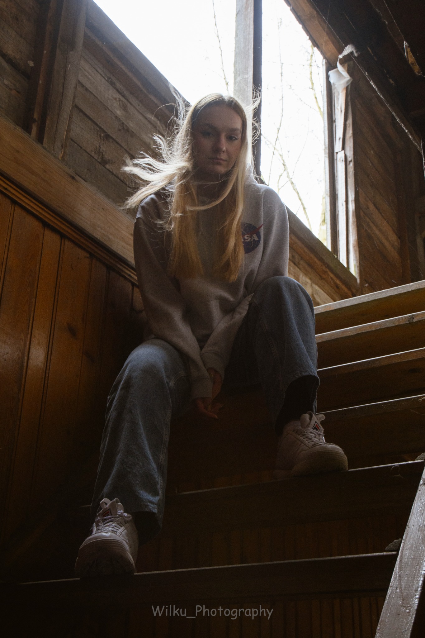 Młoda kobieta w bluzie NASA siedzi na starych drewnianych schodach w opuszczonym budynku, z światłem wpadającym przez okno w tle.