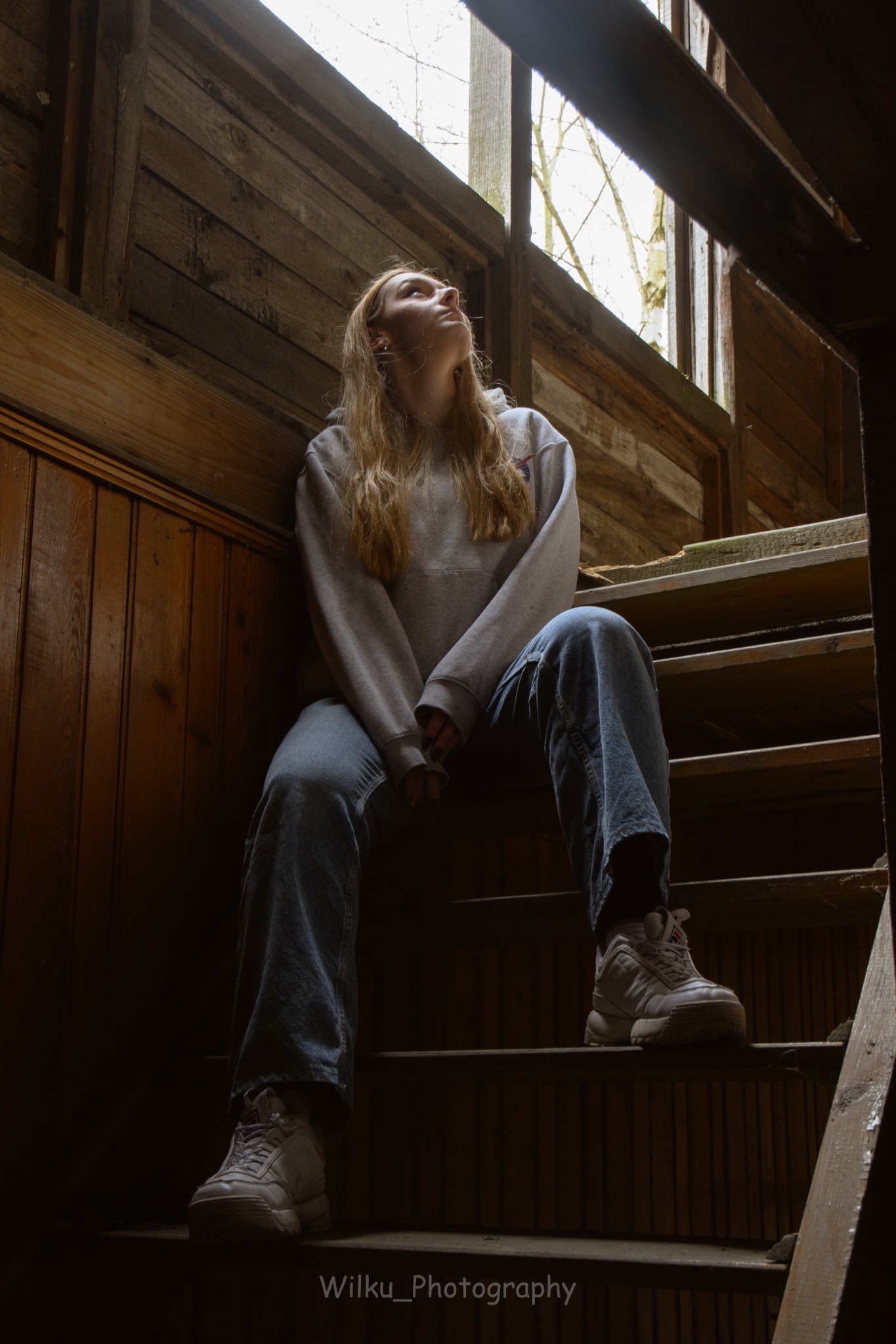 Młoda kobieta w bluzie i jeansach siedzi na drewnianych schodach w opuszczonym budynku, spoglądając w górę w stronę okna.