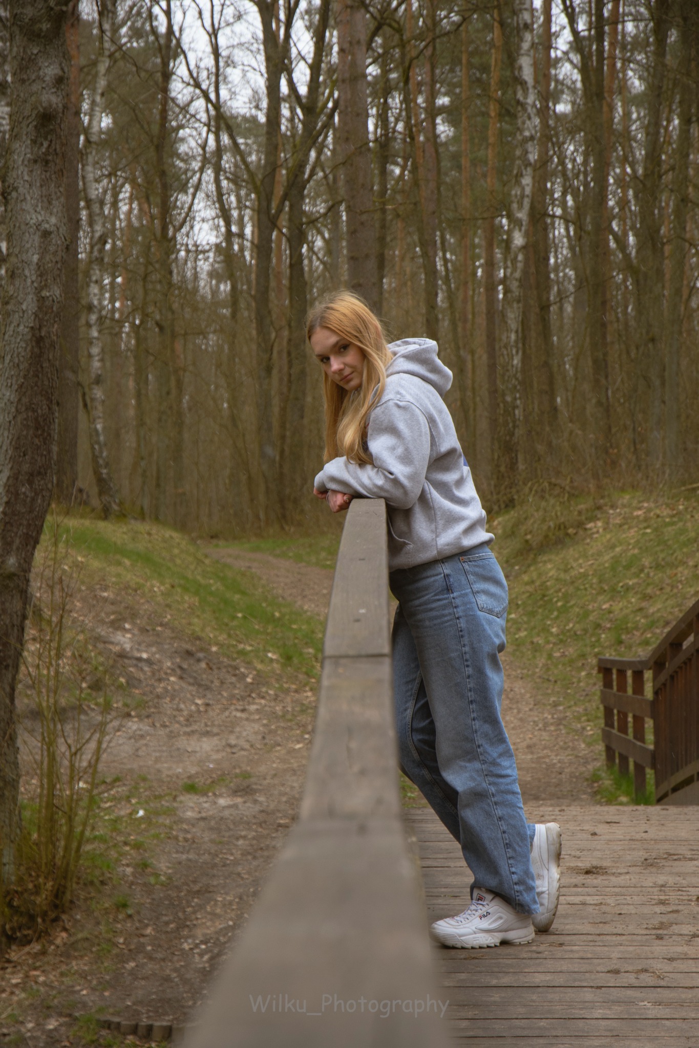 Młoda kobieta w szarej bluzie z kapturem i jeansach opiera się o drewnianą barierkę mostku w lesie, spoglądając w obiektyw.