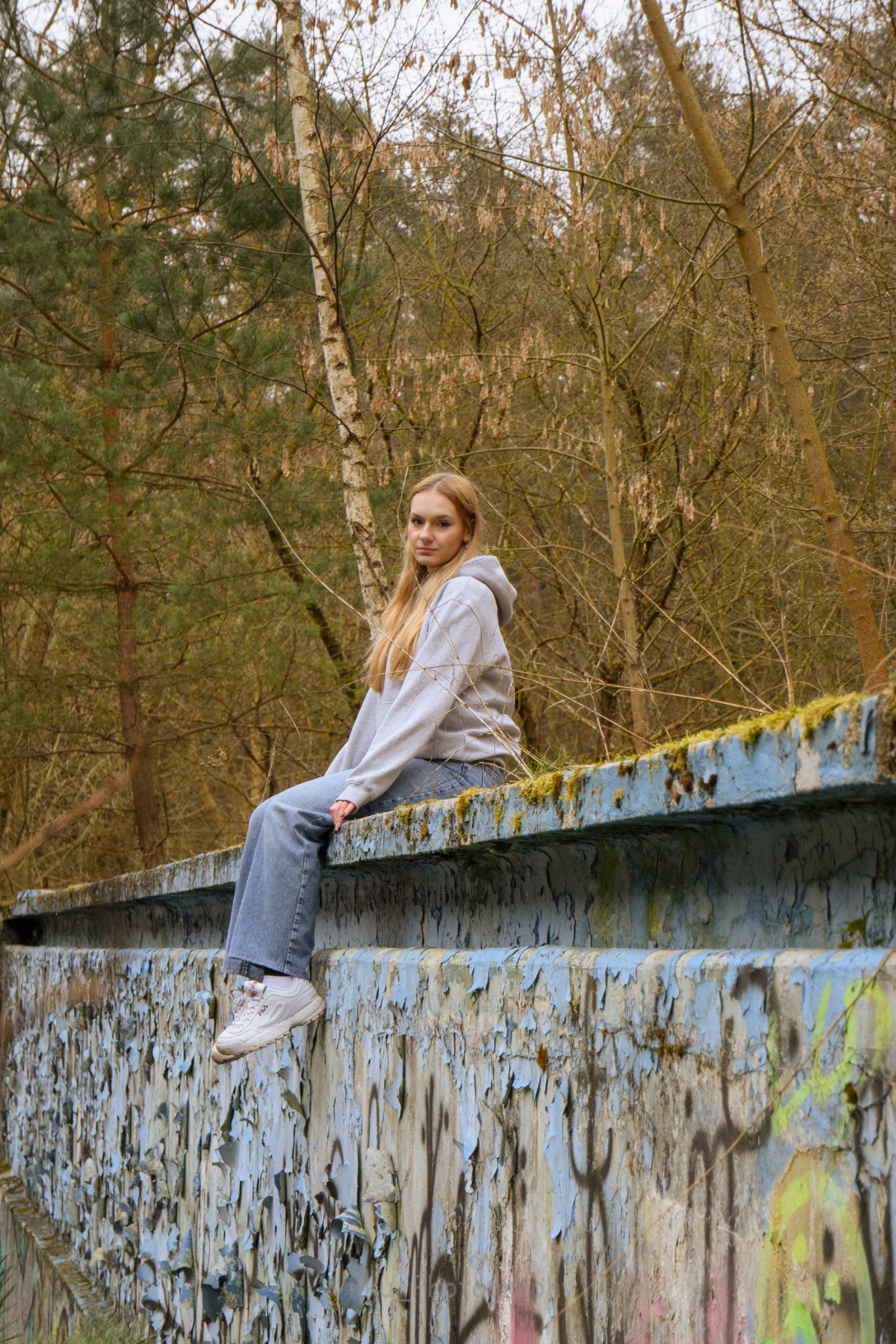 Młoda kobieta w bluzie z kapturem i dżinsach siedzi na pomalowanej na niebiesko, pokrytej mchem betonowej konstrukcji na tle drzew bez liści.
