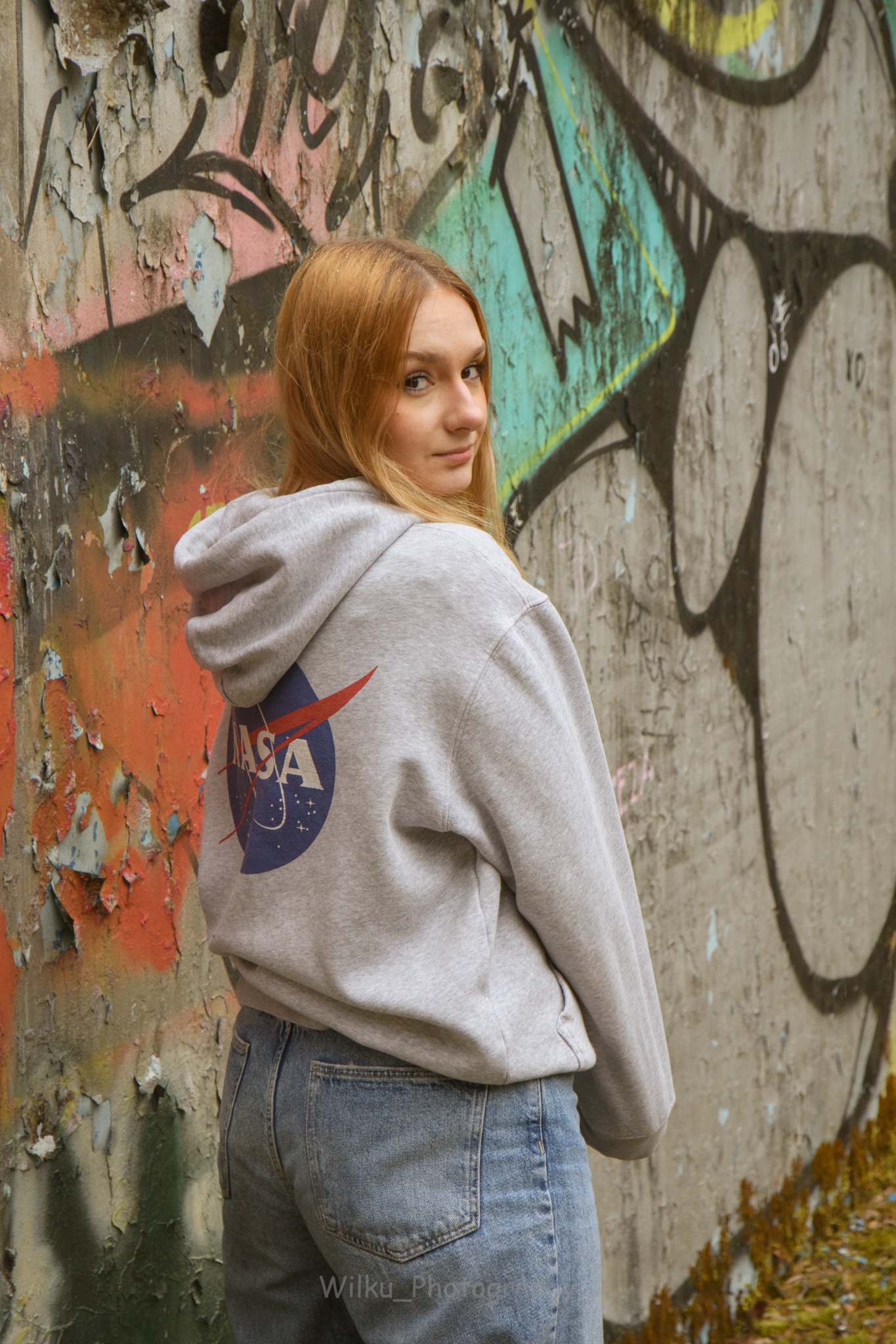 Portret kobiety w bluzie z logo NASA, odwróconej przez ramię na tle ściany pokrytej graffiti i łuszczącą się farbą.