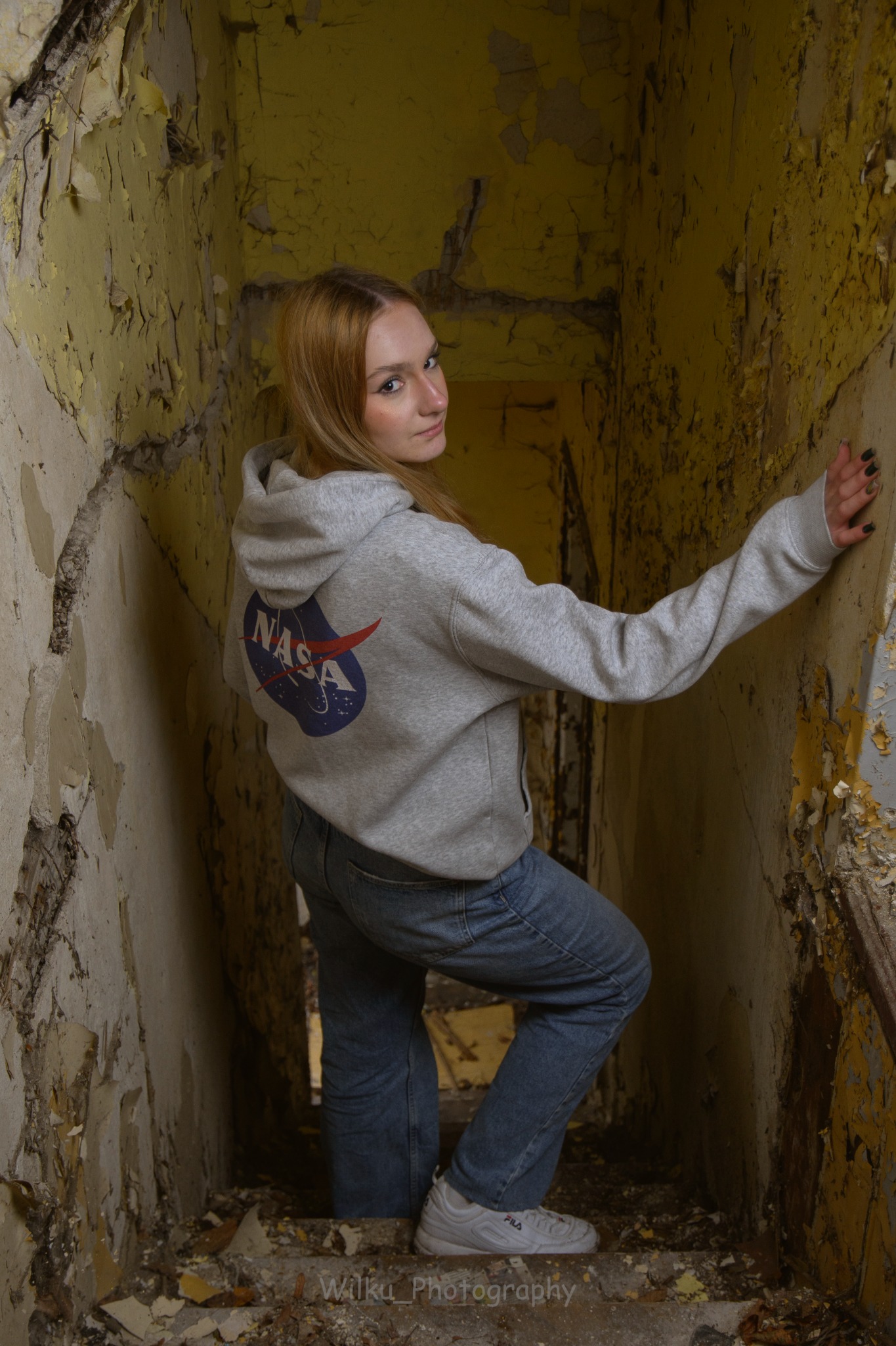Portret kobiety w bluzie z logo NASA, pozującej na zniszczonych schodach w opuszczonym budynku z odpadającą farbą.