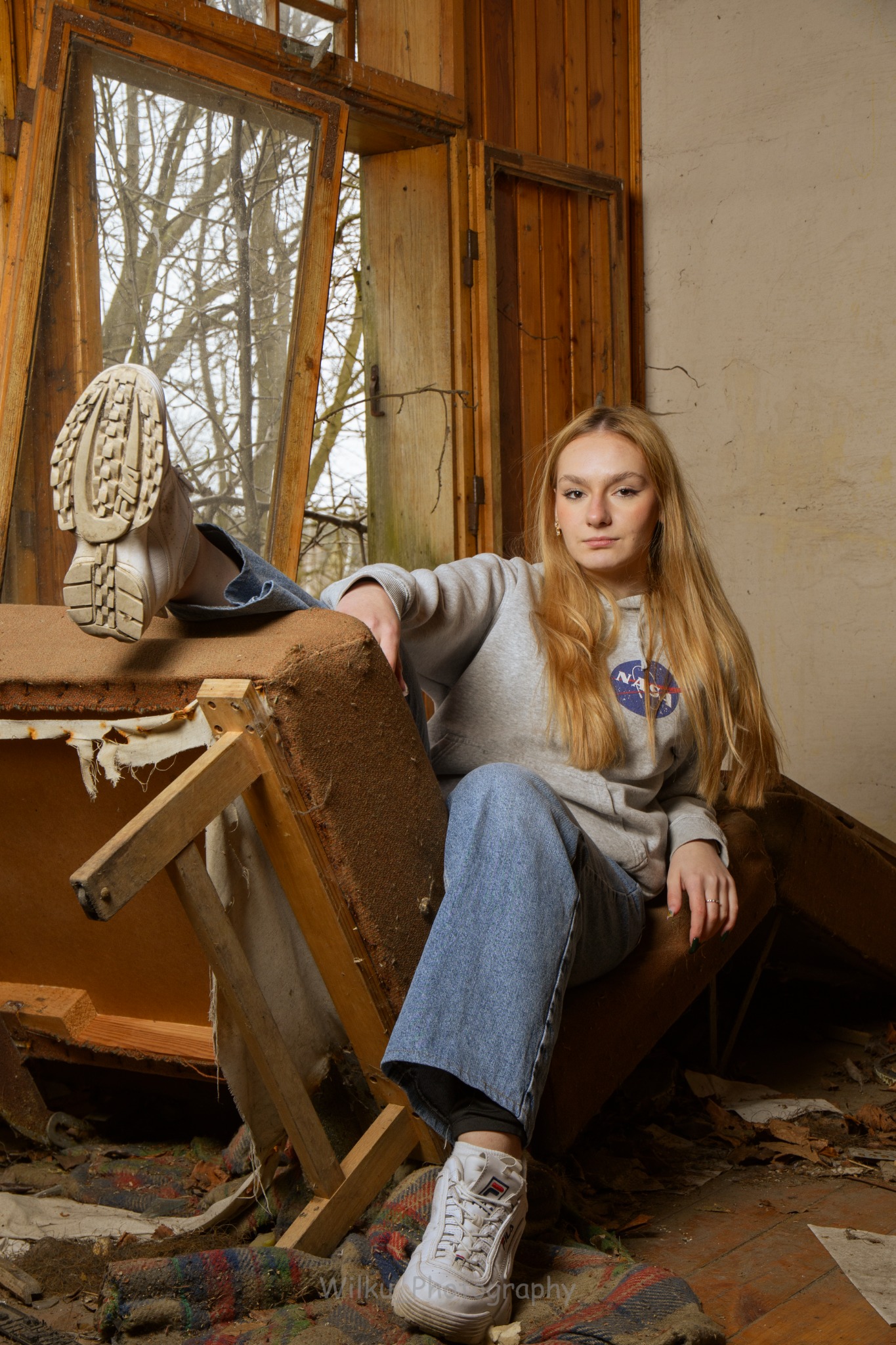Młoda kobieta w bluzie NASA i jeansach siedzi na rozpadającym się fotelu w opuszczonym pokoju z drewnianym oknem, eksponując podeszwę buta w kierunku okna.
