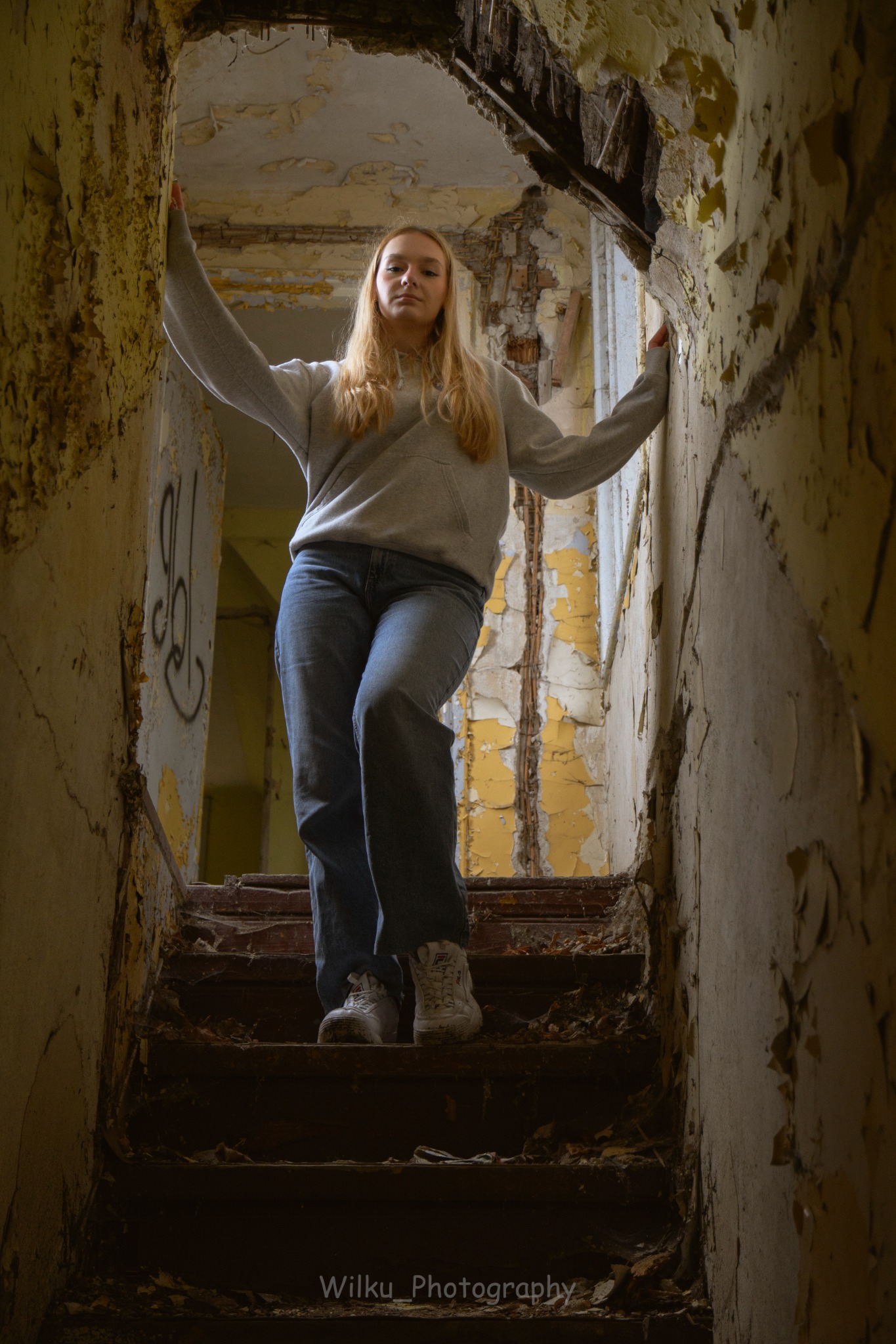 Młoda kobieta w bluzie i jeansach pozuje na zniszczonych schodach w opuszczonym budynku w Zgierzu, z widocznymi odpadającym tynkiem i graffiti.