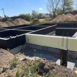 Zbrojone ławy fundamentowe z izolacją termiczną i przeciwwilgociową, widok na wykop z żółtymi kwiatami na pierwszym planie.