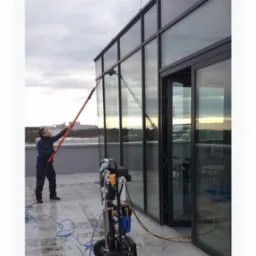 Pracownik myje szklaną elewację budynku za pomocą teleskopowej lancy i mobilnego systemu filtracji wody na dachu, widoczna różnica czystości na umytej i nieumytych częściach okna.