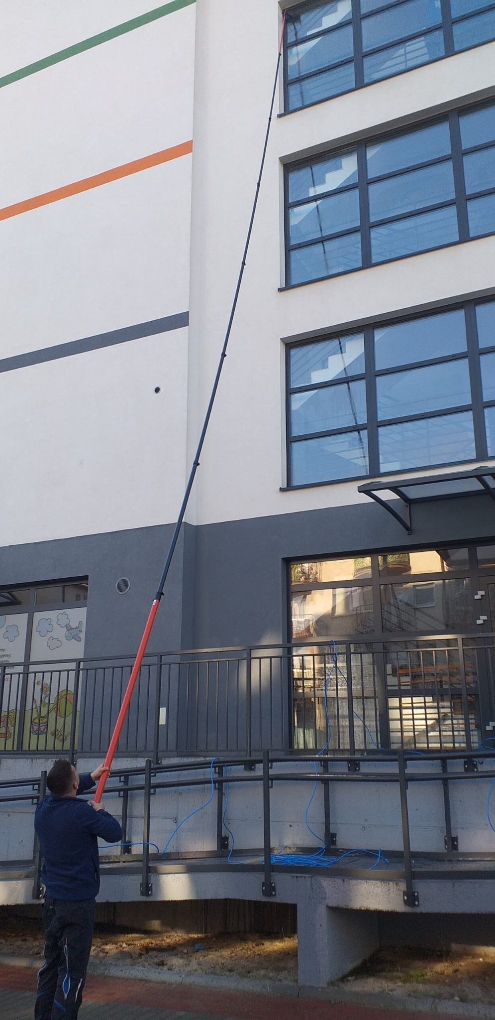 Pracownik używa teleskopowego kija z końcówką do mycia okien na wysokości w budynku z szarymi ramami okiennymi i kolorowymi pasami na elewacji.