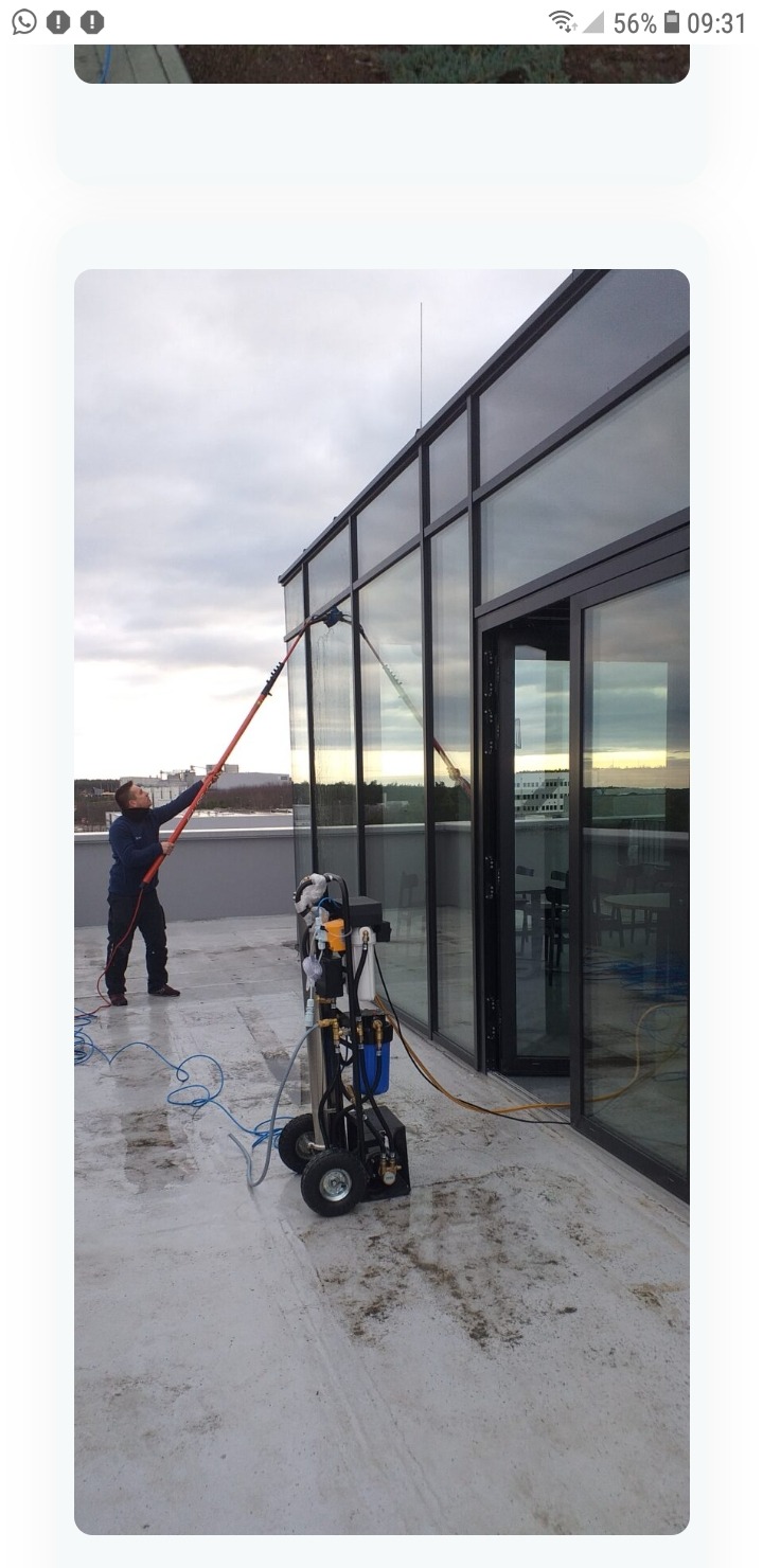 Pracownik myje szklaną elewację budynku za pomocą teleskopowej lancy i mobilnego systemu filtracji wody na dachu, widoczna różnica czystości na umytej i nieumytych częściach okna.