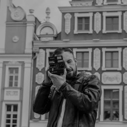 Bsshoots - Mężczyzna z brodą w skórzanej kurtce robi zdjęcie aparatem Polaroid Spirit 600 CL na tle zabytkowej architektury w czerni i bieli.