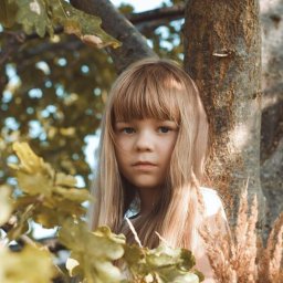 Bsshoots - Portret dziewczynki z blond włosami i grzywką na tle drzewa i liści w ciepłych, naturalnych barwach.