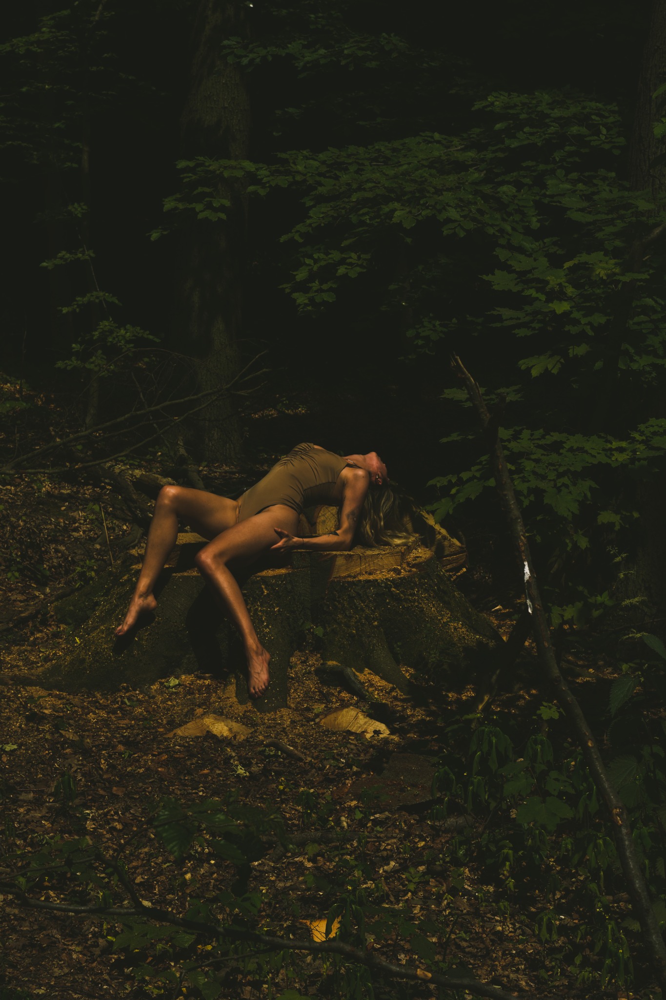 Kobieta w body leżąca na pniu drzewa w lesie, z głową odchyloną do tyłu, otoczona liśćmi i gałęziami, w naturalnym świetle.