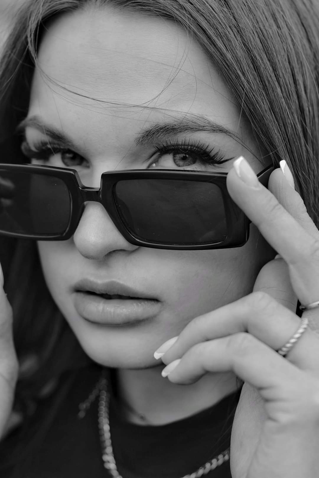 Czarno-białe portretowe ujęcie kobiety z wyrazistym makijażem oczu, trzymającej czarne okulary przeciwsłoneczne przy twarzy, widoczny srebrny łańcuszek na szyi i pierścionek na palcu.