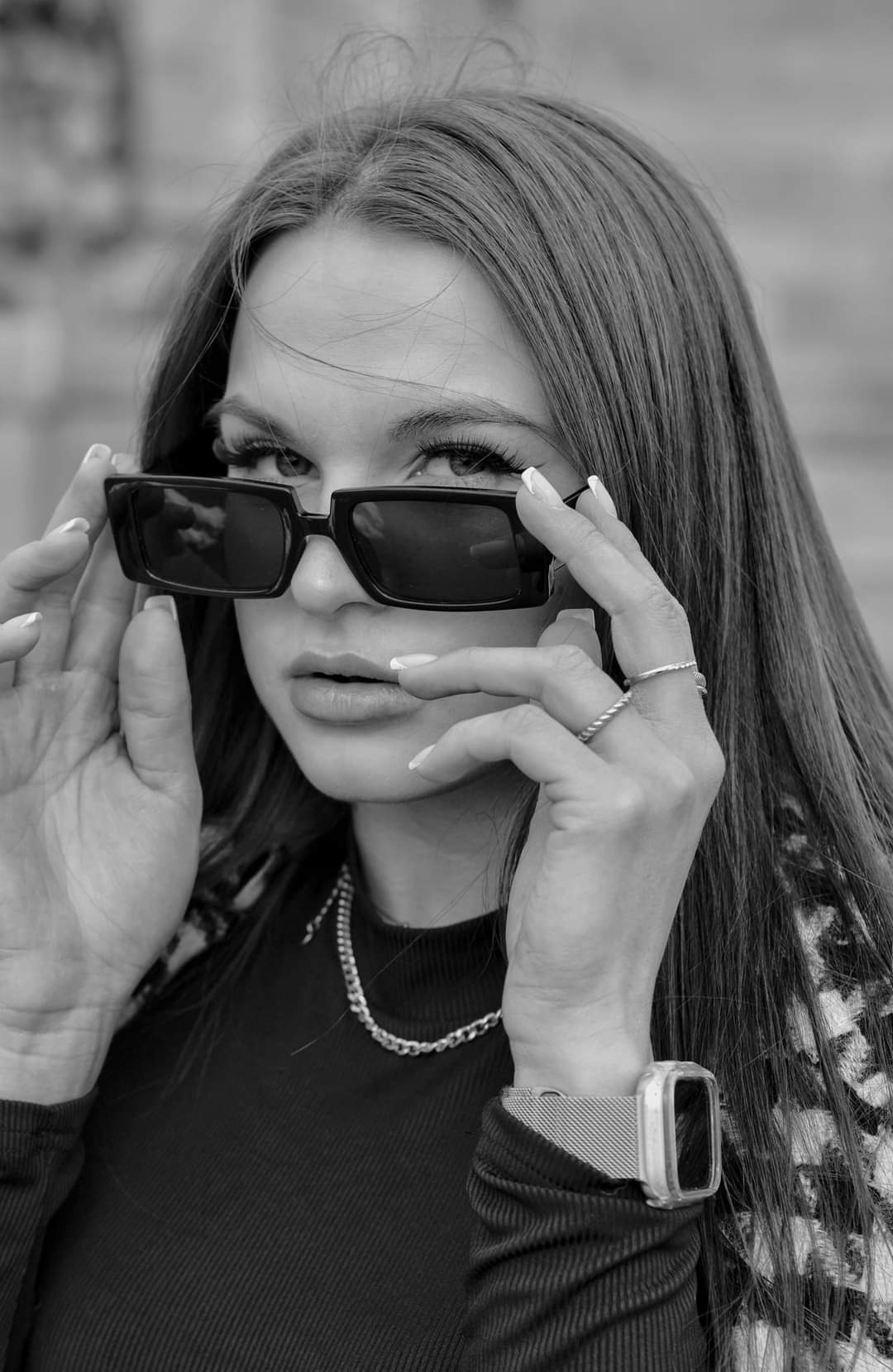 Portret młodej kobiety w okularach przeciwsłonecznych, z łańcuszkiem na szyi i zegarkiem na ręce, wykonany w czerni i bieli.