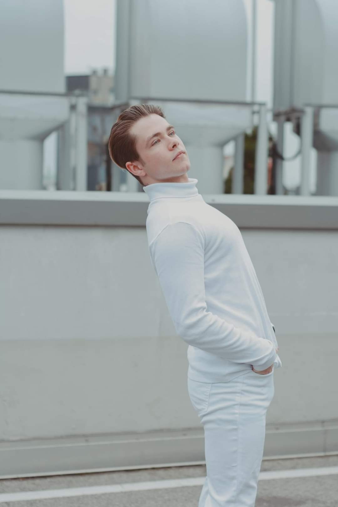 Młody mężczyzna w białym golfie i spodniach, pochylony do tyłu, z rękami w kieszeniach, spogląda w górę na tle szarej ściany i instalacji na dachu.