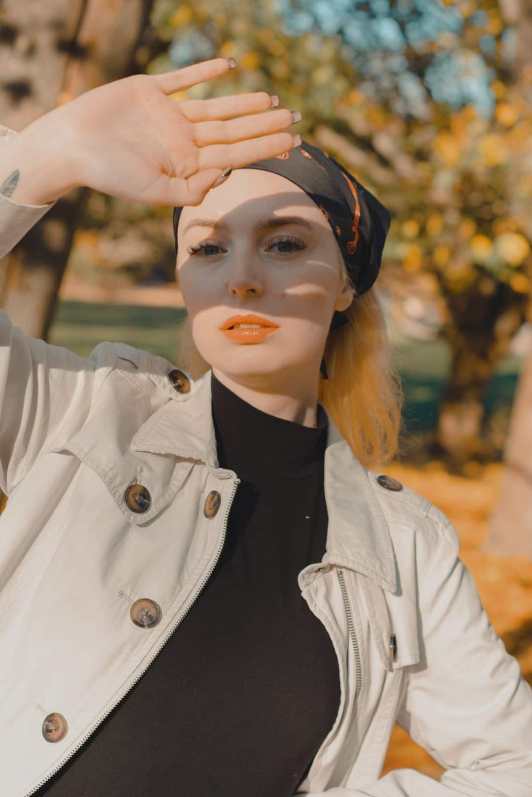 Portret kobiety w jesiennej scenerii, z dłonią osłaniającą oczy, w kremowym płaszczu i czarnym golfie, z chustą na głowie i pomarańczowymi ustami.