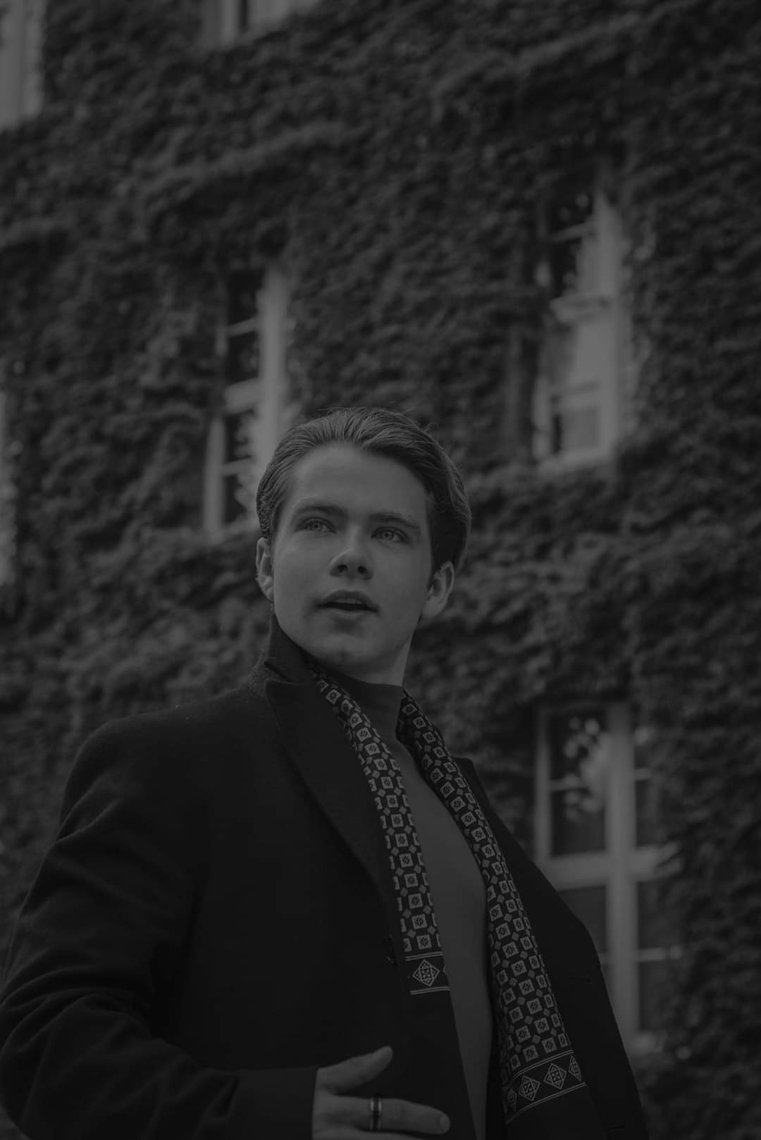 Czarno-białe portretowe ujęcie młodego mężczyzny w płaszczu i wzorzystym szaliku, spoglądającego w górę na tle budynku porośniętego bluszczem.