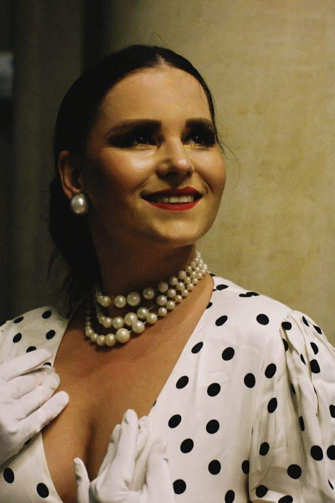 Uśmiechnięta kobieta w perłowej biżuterii i białej sukience w czarne kropki, pozująca w białych rękawiczkach.