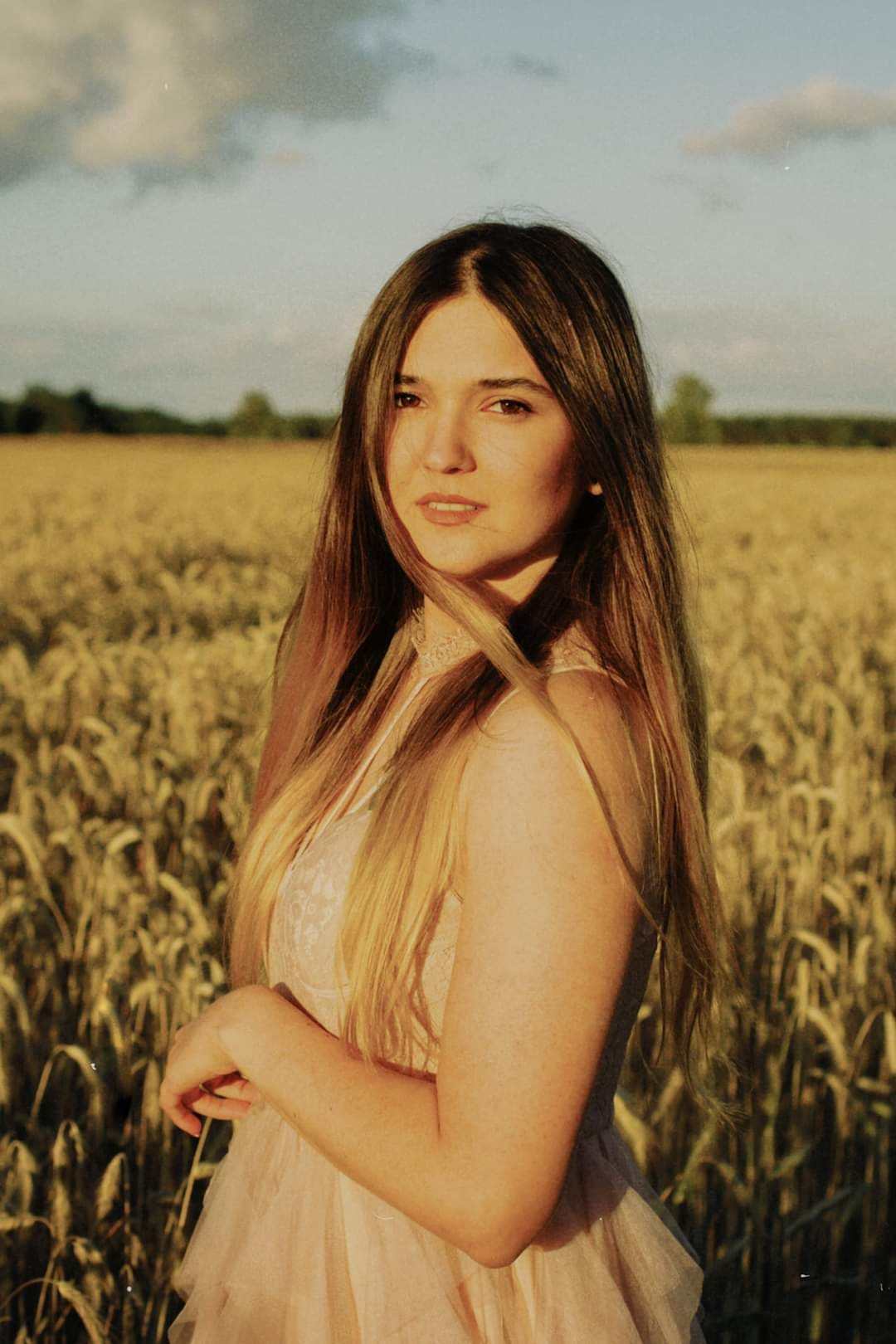 Portret młodej kobiety w zwiewnej sukience na tle pola zboża w ciepłym świetle zachodzącego słońca.