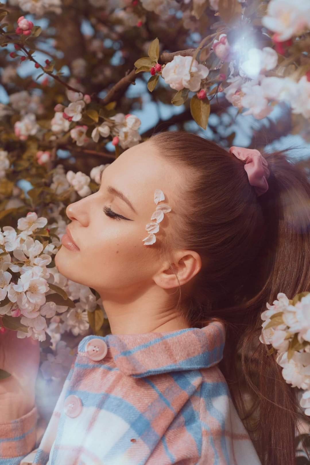 Portret kobiety z profilu, z zamkniętymi oczami, z płatkami kwiatów na twarzy, na tle kwitnącego drzewa owocowego i błękitnego nieba.