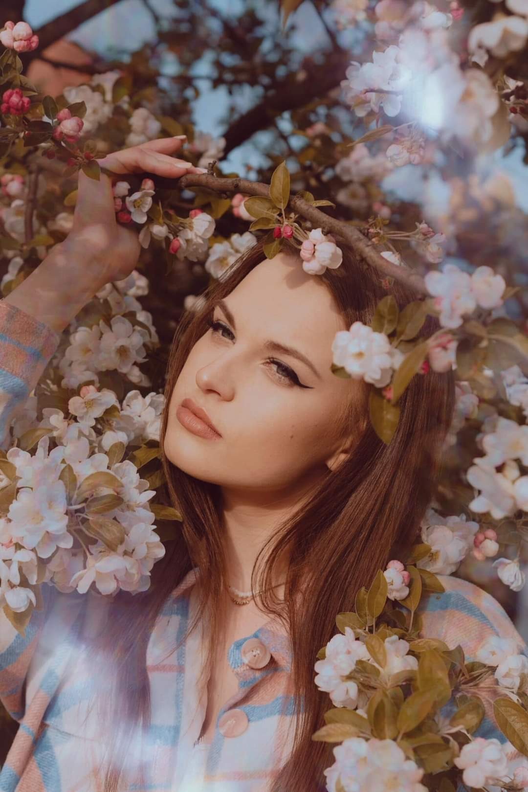 Portret młodej kobiety z długimi, brązowymi włosami, otoczonej kwitnącymi gałązkami drzewa owocowego, spoglądającej w bok z delikatnym makijażem oczu.