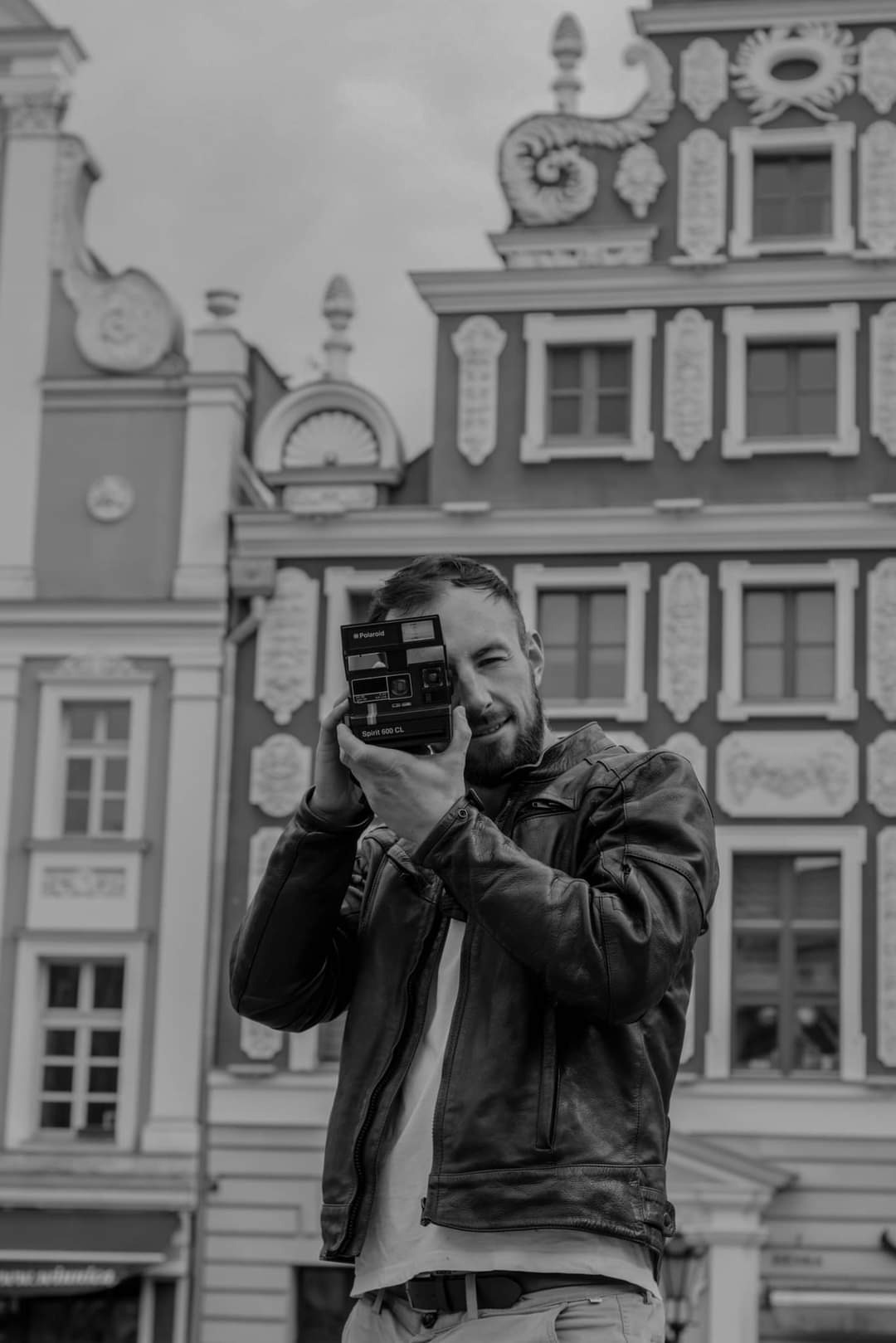 Mężczyzna z brodą w skórzanej kurtce robi zdjęcie aparatem Polaroid Spirit 600 CL na tle zabytkowej architektury w czerni i bieli.
