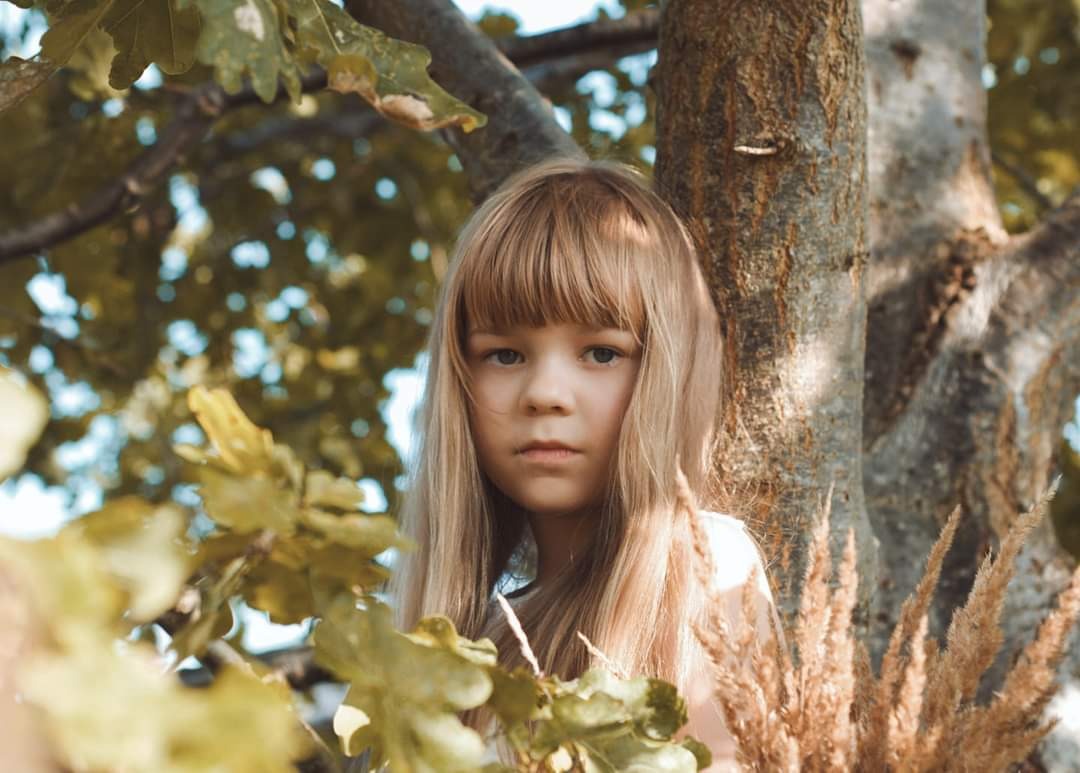 Portret dziewczynki z blond włosami i grzywką na tle drzewa i liści w ciepłych, naturalnych barwach.