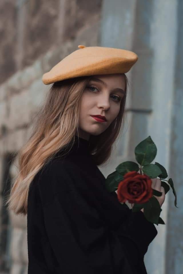 Portret młodej kobiety w czarnym golfie i żółtym berecie, trzymającej czerwoną różę na tle kamiennej ściany.