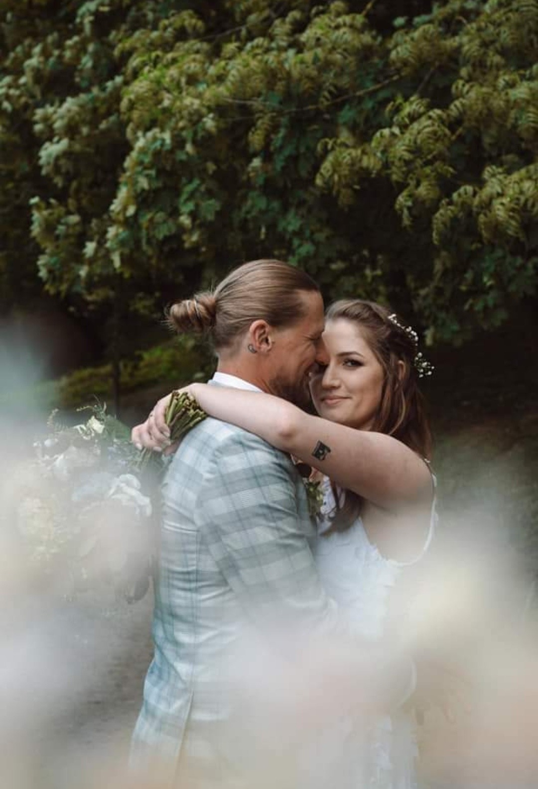 Uśmiechnięta para narzeczonych w objęciach na tle zieleni, mężczyzna w kraciastej marynarce, kobieta z kwiatami we włosach i tatuażem na ramieniu.