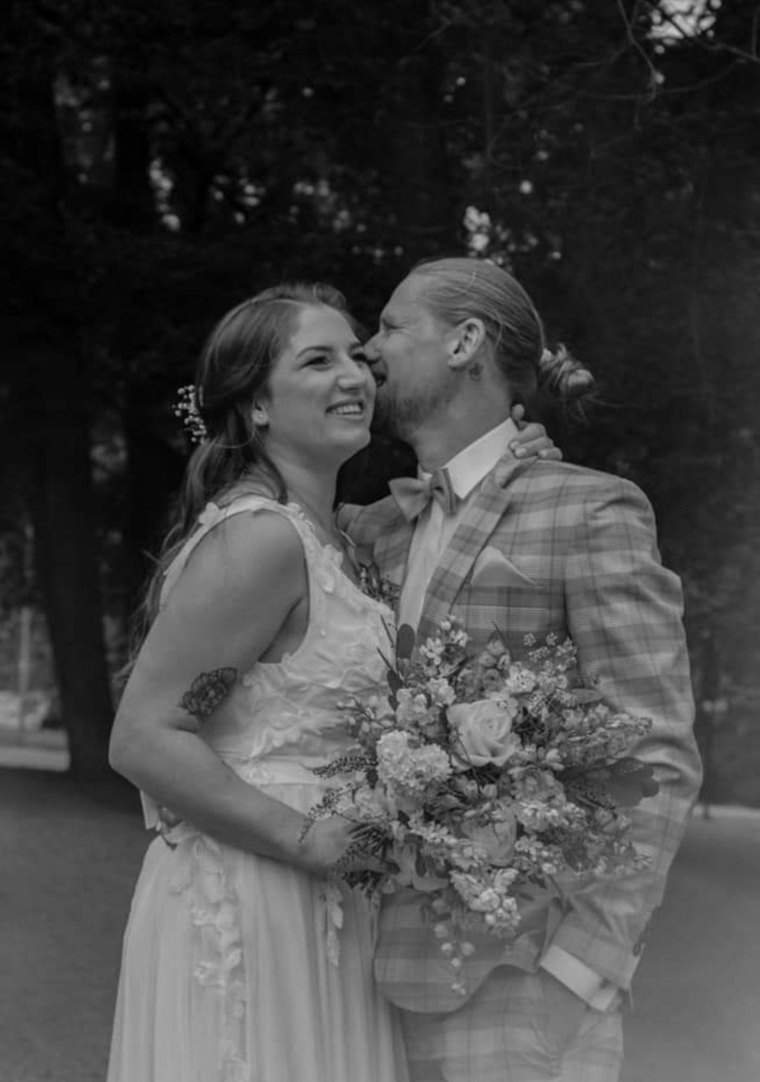 Czarno-białe zdjęcie uśmiechniętej panny młodej z bukietem, obejmowanej przez pana młodego w kraciastym garniturze z muszką.