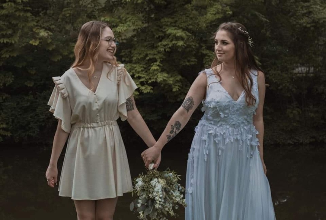 Dwie uśmiechnięte kobiety w sukienkach, trzymające się za ręce i bukiet białych kwiatów, z tatuażami na ramionach, na tle zieleni.