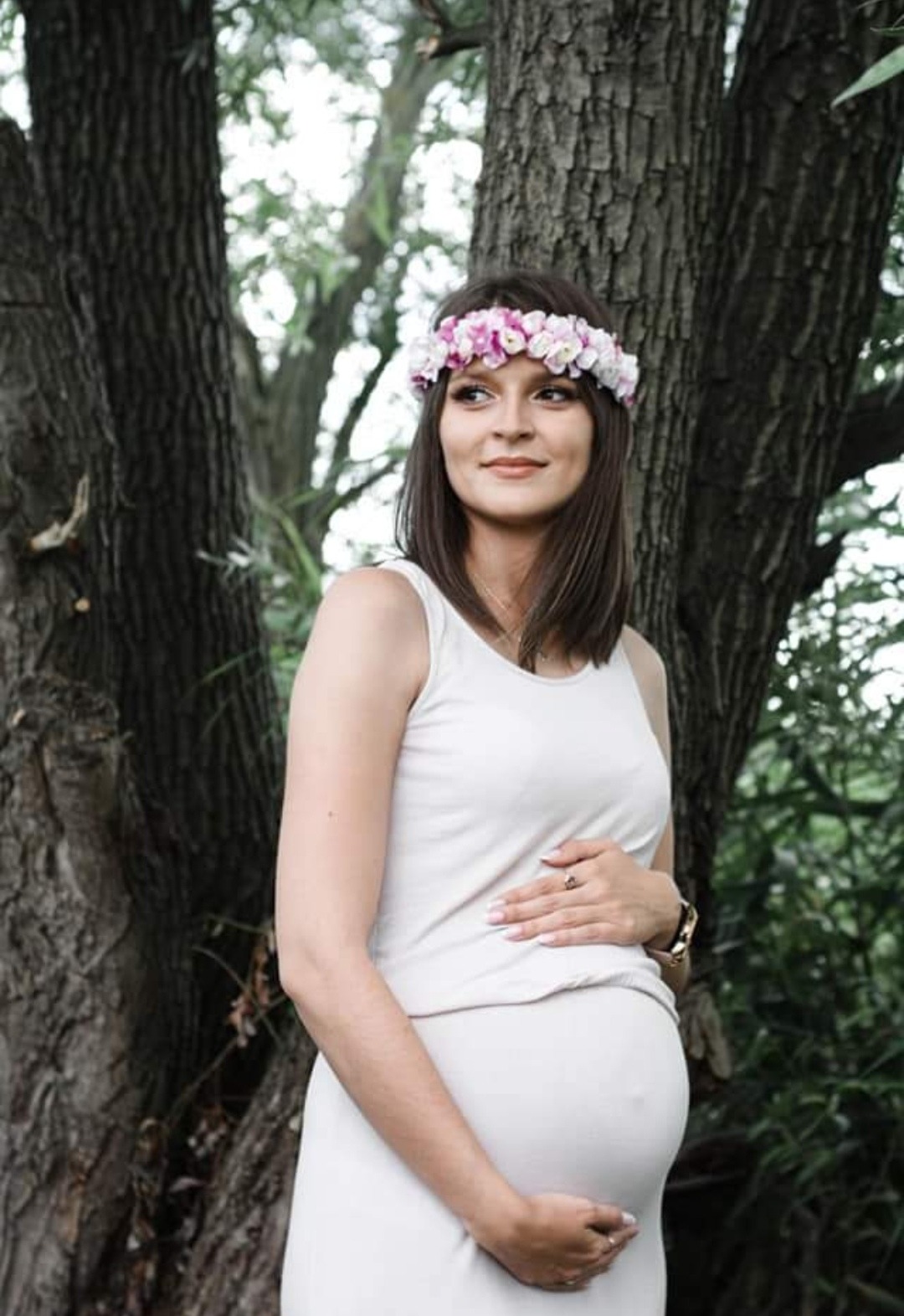 Portret kobiety w ciąży, w białej sukience i wianku z różowych kwiatów na głowie, pozującej na tle drzewa z fakturą kory.