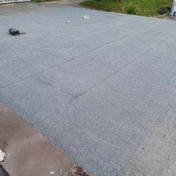 Dach-Bud Paweł Olczak - Szary dach pokryty papą termozgrzewalną, widoczne łączenia i rolka papy oraz narzędzie dekarskie, w tle budynek z blachy falistej.