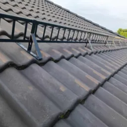 Szara dachówka ceramiczna z zamontowaną, metalową ławą kominiarską w kolorze ciemnozielonym, zabezpieczającą dostęp do komina.