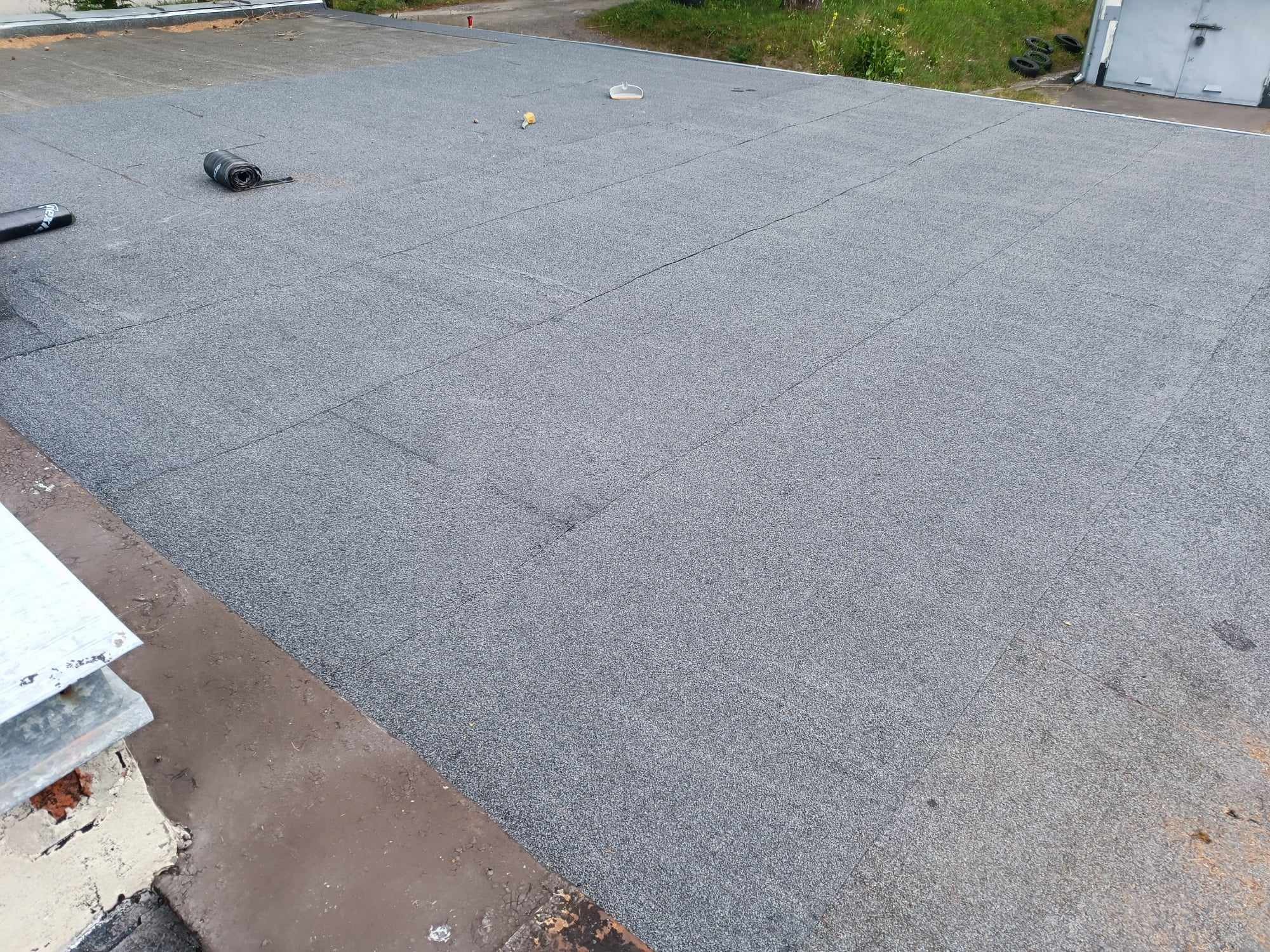 Szary dach pokryty papą termozgrzewalną, widoczne łączenia i rolka papy oraz narzędzie dekarskie, w tle budynek z blachy falistej.