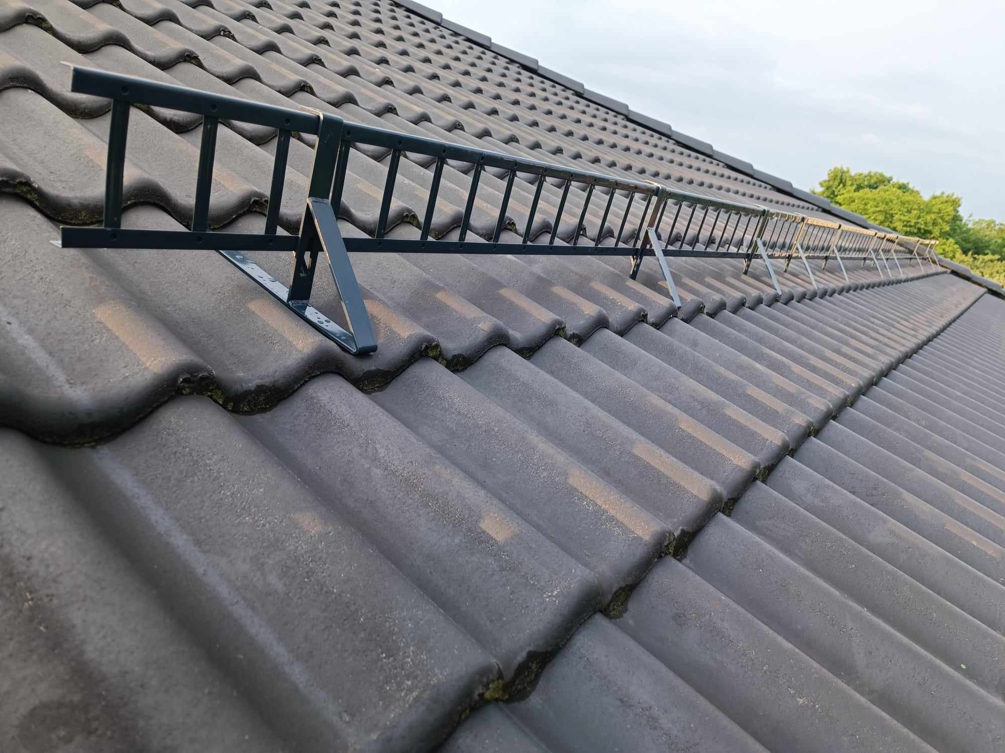 Szara dachówka ceramiczna z zamontowaną, metalową ławą kominiarską w kolorze ciemnozielonym, zabezpieczającą dostęp do komina.