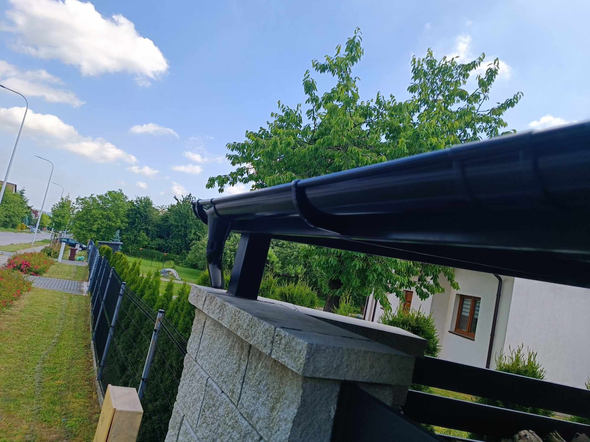 Czarny system rynnowy zamontowany na kamiennym słupku ogrodzenia, z widokiem na zielony ogród, biały dom i błękitne niebo z chmurami.