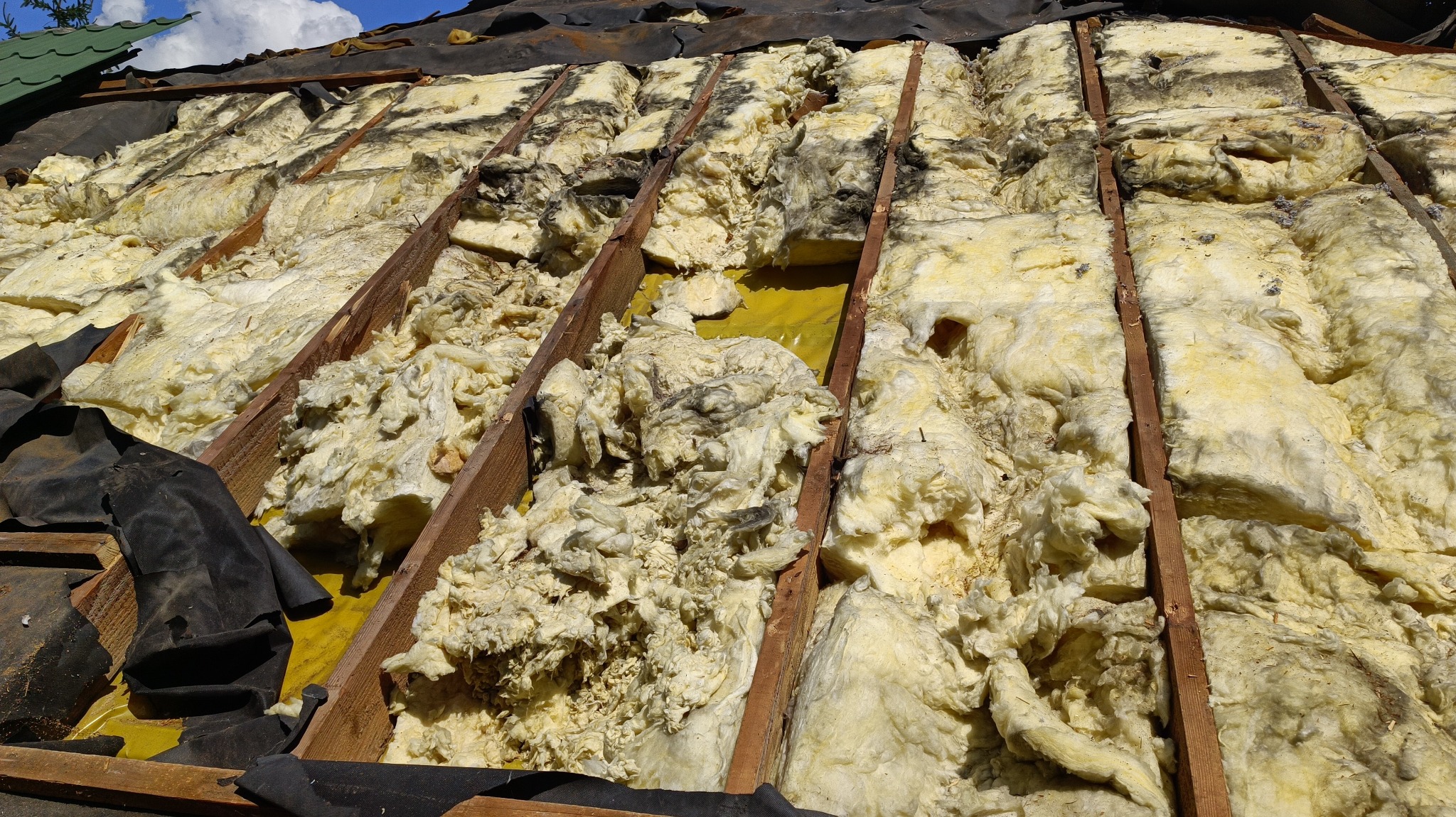 Poddasze w trakcie ocieplania, widoczne krokwie i żółta folia paroizolacyjna, pomiędzy nimi wełna mineralna z widocznymi zabrudzeniami i miejscowymi ubytkami, czarna membrana dachowa przy krawędzi.