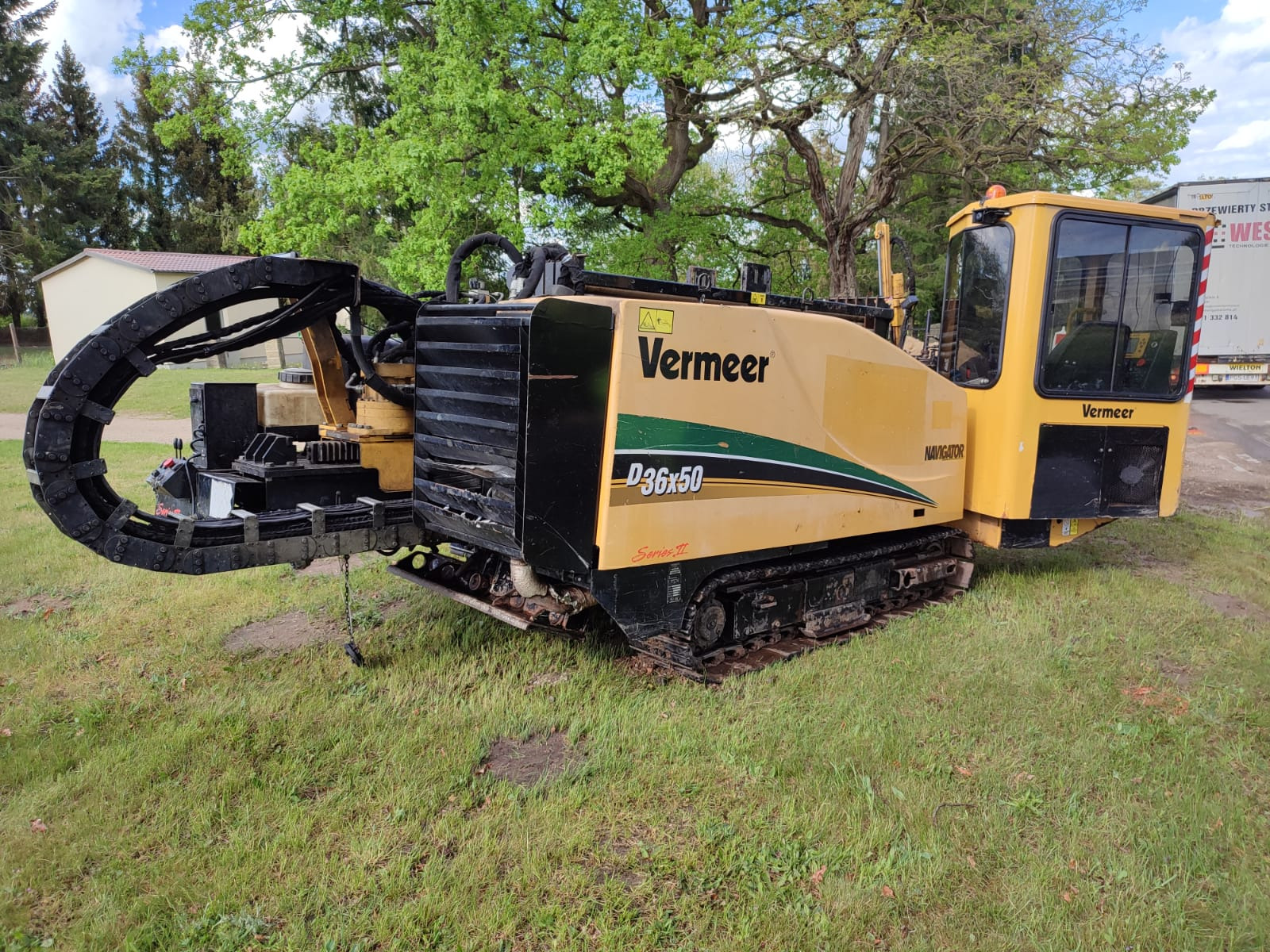 Żółta maszyna Vermeer D36x50 Navigator do wiercenia horyzontalnego na gąsienicach, zaparkowana na trawie, z widocznym łańcuchem podajnika wiertła, z tyłu widoczny fragment ciężarówki.