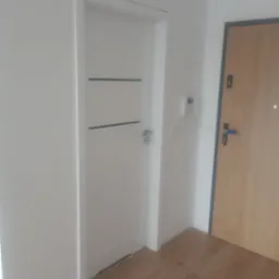 Białe drzwi wewnętrzne z poziomymi, ciemnymi paskami, zamontowane w białej ścianie obok drewnianych drzwi wejściowych i domofonu, na podłodze z paneli.