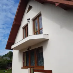 Świeżo wykończona elewacja domu jednorodzinnego z brązowym dachem i drewnianą stolarką okienną, widok z dołu, na trawniku widoczne wiadro po farbie elewacyjnej.