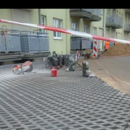 Widok na plac z ułożoną ażurową kostką betonową, piła do cięcia betonu, ubijarka, kanister i inne narzędzia budowlane w tle, robotnicy w pomarańczowych kamizelkach przy pryzmie piachu.