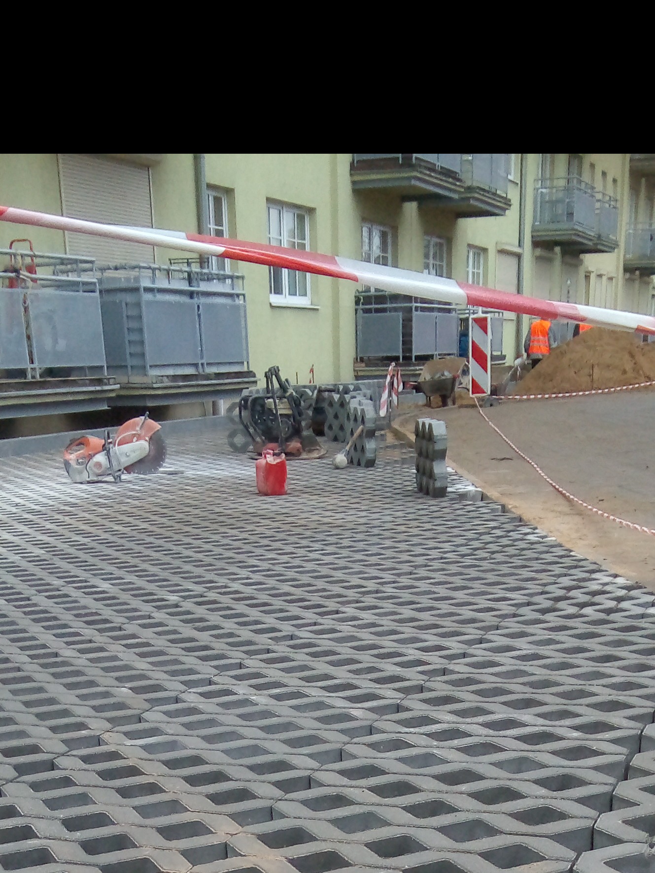 Widok na plac z ułożoną ażurową kostką betonową, piła do cięcia betonu, ubijarka, kanister i inne narzędzia budowlane w tle, robotnicy w pomarańczowych kamizelkach przy pryzmie piachu.