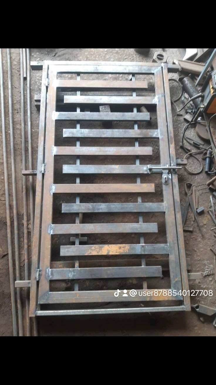 Surowa, metalowa brama z poziomymi szczeblami i prostym zamknięciem, widoczna w warsztacie obok narzędzi i prętów, jeszcze przed malowaniem.