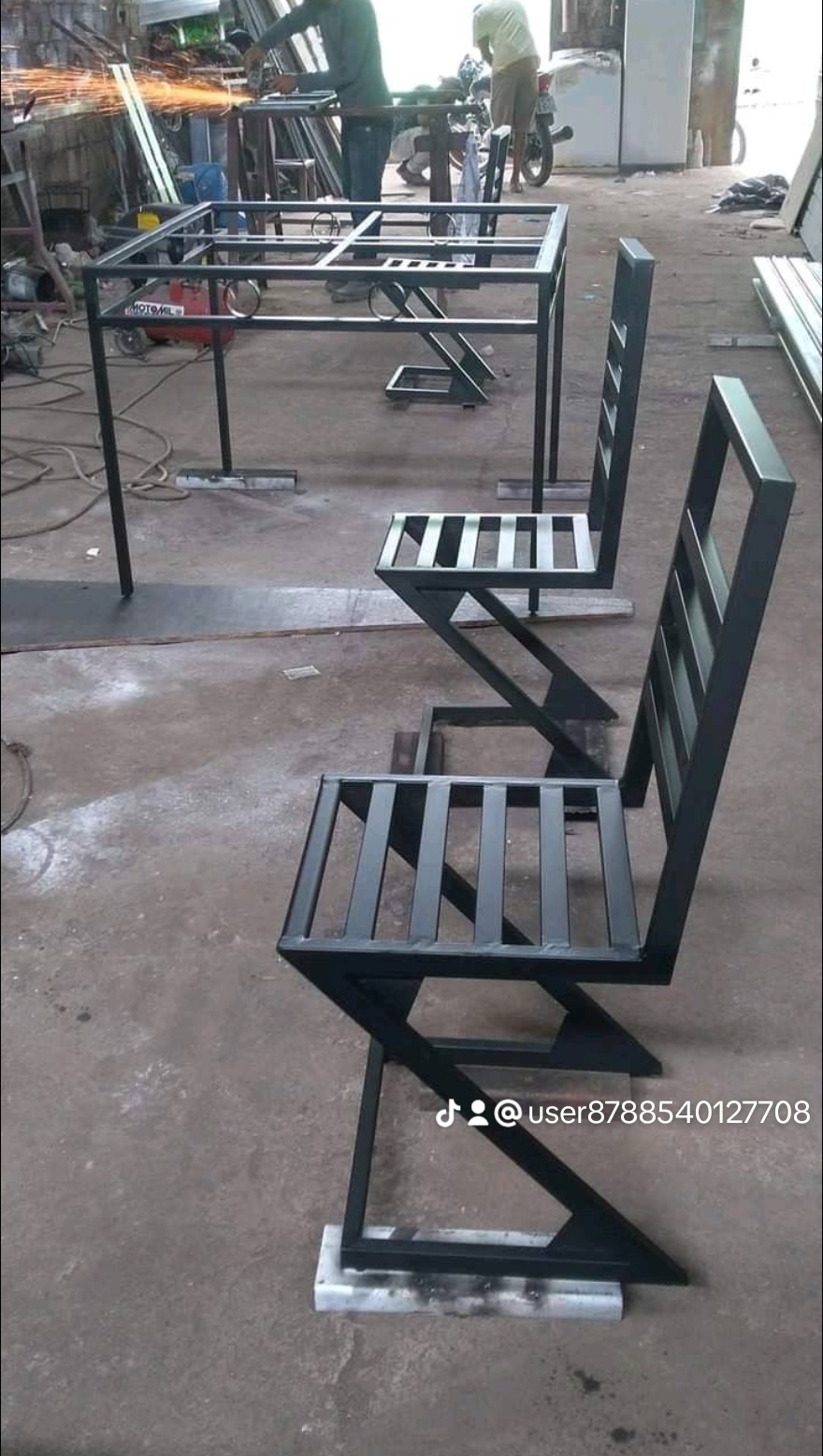 Metalowa konstrukcja stołu i dwóch krzeseł o geometrycznym kształcie, spawana w warsztacie, widoczne iskry podczas obróbki.