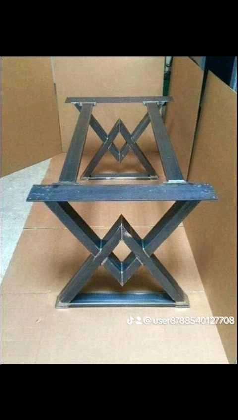 Dwie surowe, metalowe podstawy stołu o geometrycznym wzorze, z widocznymi spawami, ustawione na kartonowym podłożu.