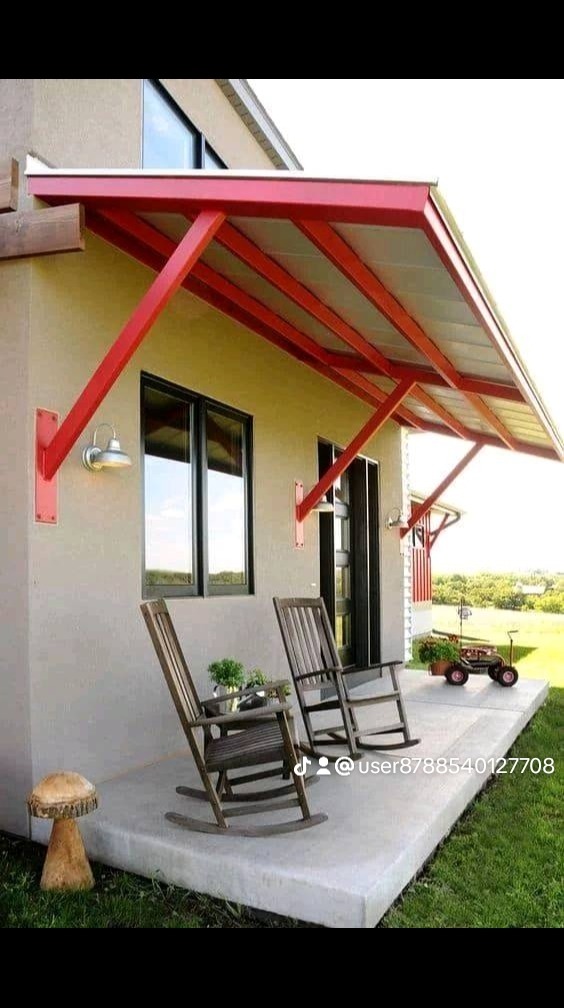 Zadaszenie nad wejściem do domu z czerwonymi elementami konstrukcyjnymi, widoczne dwa drewniane fotele bujane na betonowym tarasie, dekoracja w kształcie grzyba i zabawkowy wózek w tle.